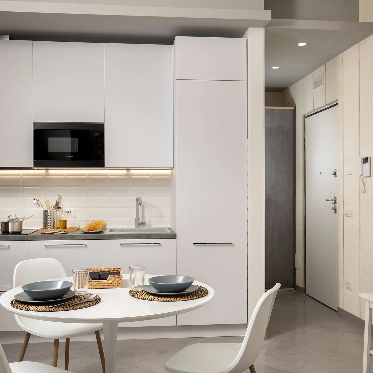 Monolocale moderno con cucina attrezzata, tavolo rotondo con piatti e sedia, accanto a una porta d’ingresso